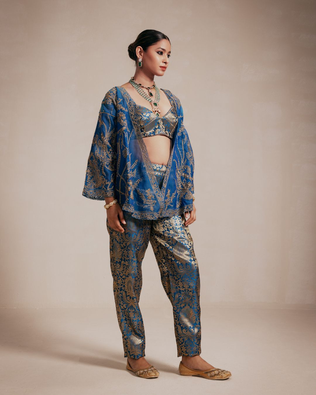 A woman models a traditional Indian outfit featuring a blue and gold patterned kurta, matching trousers, a sheer mesh overlay, and a matching embellished crop top. She is standing against a solid beige studio backdrop.