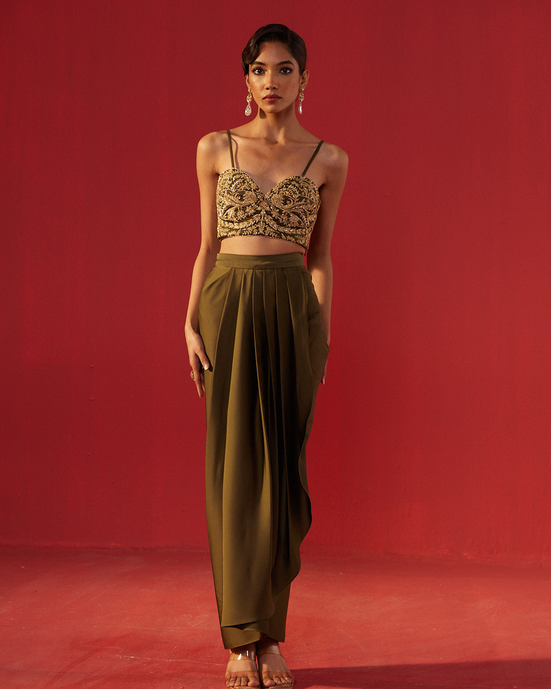 A model stands against a solid red studio background wearing a two-piece outfit consisting of a gold heavily-embroidered crop top with thin black straps and high-waisted olive green draped maxi pants.